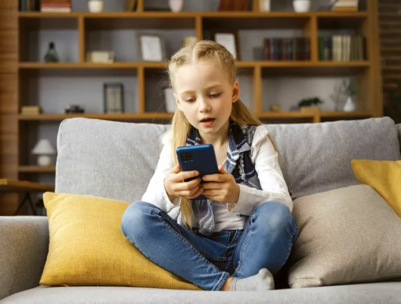 Portrait of cute little cute girl holding smartphone and resting at sofa at home. Choosing favorite music or cartoons,texting messages,browsing internet,watching video, playing games on mobile phone Jak smartfony zmieniły dzieciństwo na gorsze –i jak to naprawić