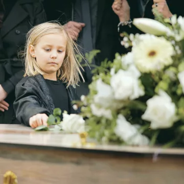 Death, grief and girl at funeral with flower on coffin, family and sad child at service in graveyard for respect. Roses, loss and people at wood casket in cemetery with kid crying at grave for burial Jak rozmawiać z dziećmi o śmierci?